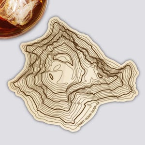 May include: A wooden coaster with a topographic map design. The coaster features contour lines in shades of brown, depicting the Charity South Sister OR mountain range. The coaster is light-colored with a natural wood grain.