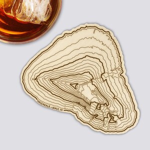 Könnte beinhalten: Ein hellfarbiger Holzuntersetzer mit einem topografischen Kartendesign. Der Untersetzer hat dunkelbraune Konturlinien und den Text "HALEAKALA • MAUI, HI". Ein Glas bernsteinfarbener Flüssigkeit mit Eis ist teilweise in der oberen linken Ecke zu sehen.