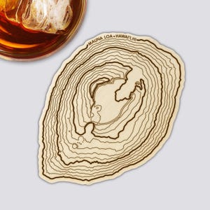 Könnte beinhalten: Ein Untersetzer aus Holz mit einem topografischen Kartendesign von Mauna Loa, Hawaii. Der Untersetzer ist hellbraun mit dunkelbraunen Konturlinien und dem Text "MAUNA LOA - HAWAII, HI". Ein Glas bernsteinfarbener Flüssigkeit ist oben links zu sehen.