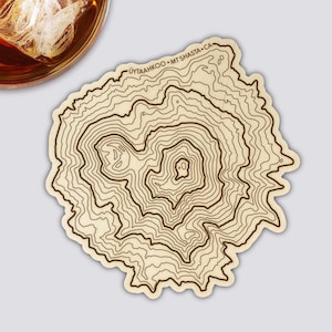 Könnte beinhalten: Ein Untersetzer aus Holz mit einem topografischen Kartendesign. Der Untersetzer hat braune Konturlinien auf einem hellbeige Hintergrund. Der Text "UYTAHKOO • MT SHASTA • CA" ist am oberen Rand aufgedruckt. Ein Glas bernsteinfarbener Flüssigkeit mit Eis befindet sich in der oberen linken Ecke.
