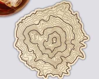May include: A wooden coaster with a topographic map design. The coaster features brown contour lines on a light beige background. The text "UYTAHKOO • MT SHASTA • CA" is printed along the top edge. A glass of amber liquid with ice is in the upper left corner.