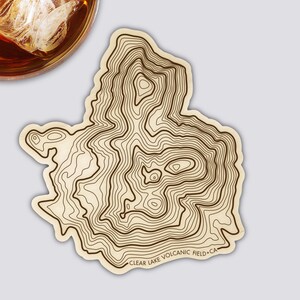Könnte beinhalten: Ein Holzuntersetzer mit einem topografischen Kartendesign. Der Untersetzer hat dunkelbraune Konturlinien auf hellbraunem Hintergrund. Der Text "CLEAR LAKE VOLCANIC FIELD CA" ist am unteren Rand aufgedruckt.