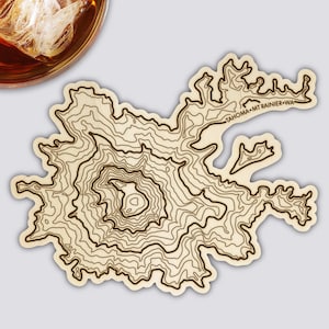 Könnte beinhalten: Ein Holzuntersetzer mit einem topografischen Kartendesign des Mount Rainier, Washington. Der Untersetzer hat dunkelbraune Konturlinien auf hellbraunem Hintergrund. Der Text "TAHOMA MT RAINIER WA" ist aufgedruckt.