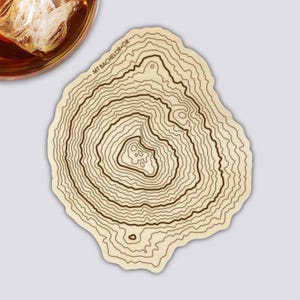 Könnte beinhalten: Ein hellfarbiger, unregelmäßig geformter Untersetzer mit einem topografischen Kartendesign. Die Karte zeigt konzentrische Linien, die die Höhe darstellen, mit dem Text "MT BACHELOR - OR" am oberen Rand. Ein Glas bernsteinfarbener Flüssigkeit mit Eis ist teilweise sichtbar.