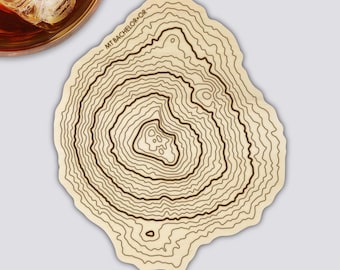 May include: A light-colored, irregularly shaped coaster with a topographic map design. The map features concentric lines representing elevation, with the text "MT BACHELOR - OR" along the top edge. A glass of amber liquid with ice is partially visible in the upper left corner.