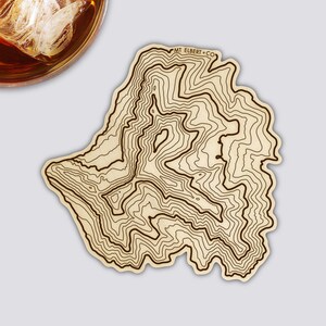 Könnte beinhalten: Ein Untersetzer aus Holz mit einem topografischen Kartendesign. Der Untersetzer ist hellbraun mit dunkelbraunen Konturlinien. Der Text "MT ELBERT CO" ist oben rechts aufgedruckt. Ein Glas bernsteinfarbener Flüssigkeit mit Eis ist oben links teilweise sichtbar.