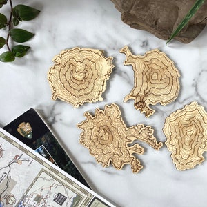 May include: Four wooden coasters featuring topographic maps of mountains. The coasters are light brown and have a natural wood finish.