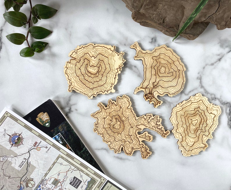 May include: Four wooden coasters featuring topographic maps of mountains. The coasters are light brown and have a natural wood finish.