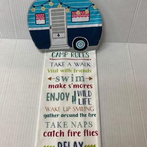 May include: A white kitchen towel with a blue and white camper van design at the top. The towel has text that reads "Camp Rules: Take a walk, Visit with friends, -swim-, make s'mores, Enjoy Wild Life, Wake up smiling, gather around the fire, Take naps, catch fire flies, Relax."