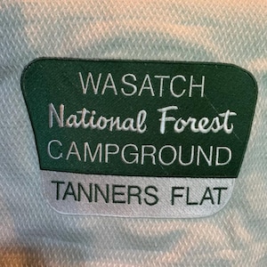 May include: A light green fabric with a dark green and white embroidered patch. The patch reads "WASATCH National Forest CAMPGROUND TANNERS FLAT" in white lettering.