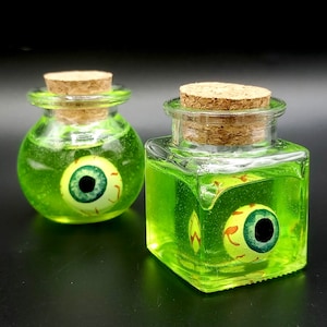 May include: Two glass jars with cork lids filled with green liquid and a floating eyeball. The jars are on a black surface.