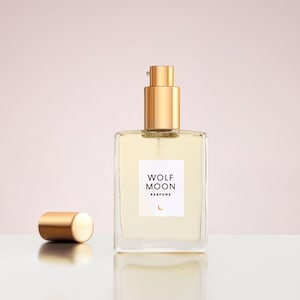 May include: A glass bottle of Wolf Moon perfume with a gold cap and a crescent moon logo. The bottle is sitting on a white surface.