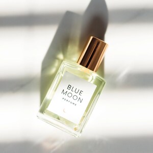K&ouml;nnte beinhalten: Eine durchsichtige Glasflasche mit Parfum und einem goldenen Verschluss. Das Etikett tr&auml;gt die Aufschrift "BLUE MOON PERFUME" mit einem Halbmond-Symbol.