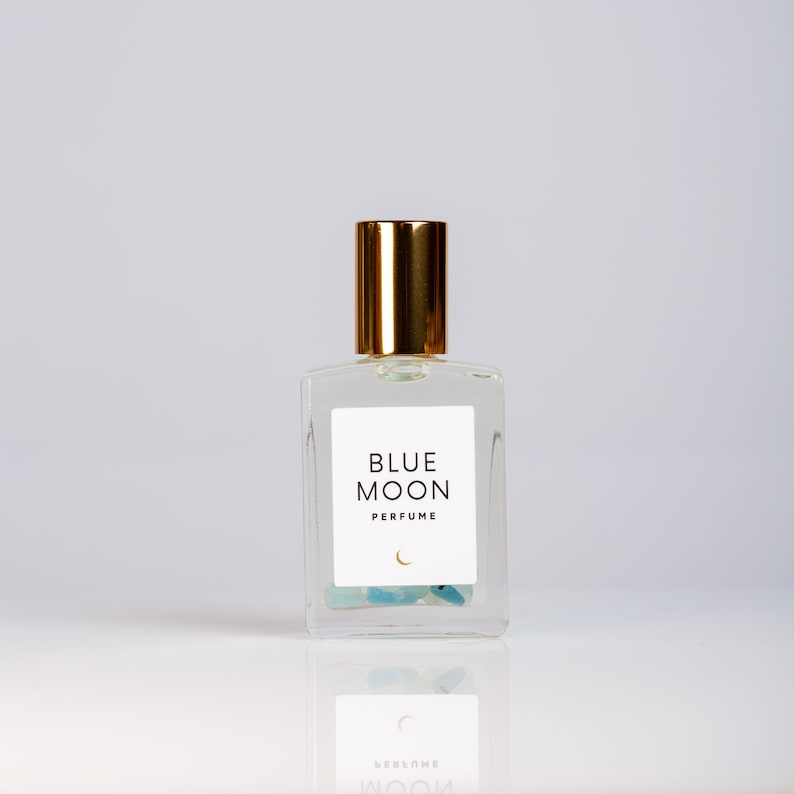 K&ouml;nnte beinhalten: Eine durchsichtige Glasflasche mit Parfum, goldener Kappe und einem wei&szlig;en Etikett mit der Aufschrift "BLUE MOON PERFUME" sowie einem Halbmond und einem Stern. Die Flasche ist mit einer klaren Fl&uuml;ssigkeit gef&uuml;llt und enth&auml;lt kleine blaue und gr&uuml;ne Steine.