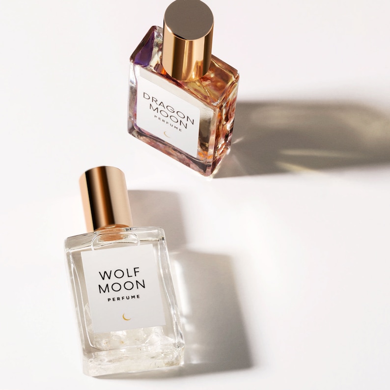 Puede incluir: Dos botellas de perfume de vidrio con tapas doradas. Las botellas est&aacute;n etiquetadas como "Dragon Moon Perfume" y "Wolf Moon Perfume" con un s&iacute;mbolo de luna creciente.
