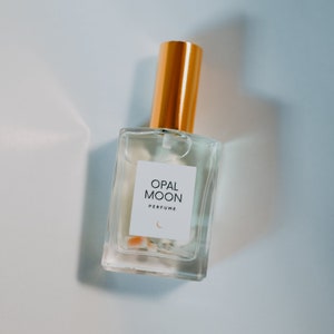 Puede incluir: Una botella de perfume de vidrio transparente con una tapa dorada. La etiqueta dice "OPAL MOON PERFUME" con un s&iacute;mbolo de luna creciente.