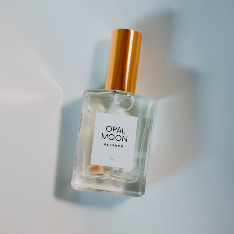 Puede incluir: Una botella de perfume de vidrio transparente con una tapa dorada. La etiqueta dice "OPAL MOON PERFUME" con un s&iacute;mbolo de luna creciente.