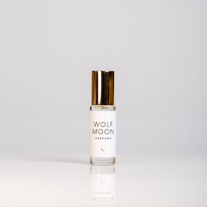 Puede incluir: Un peque&ntilde;o frasco de vidrio de perfume Wolf Moon con una tapa dorada. La etiqueta es blanca con texto negro que dice "Wolf Moon Perfume" y un s&iacute;mbolo de luna creciente.