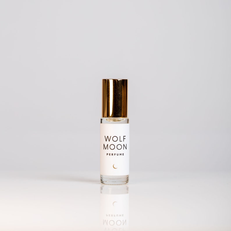 Puede incluir: Un peque&ntilde;o frasco de vidrio de perfume Wolf Moon con una tapa dorada. La etiqueta es blanca con texto negro que dice "Wolf Moon Perfume" y un s&iacute;mbolo de luna creciente.