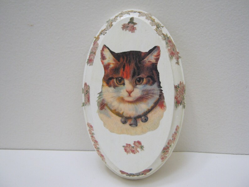 May include: White oval wooden plaque with a floral border and a vintage image of a cat with a bell collar.
