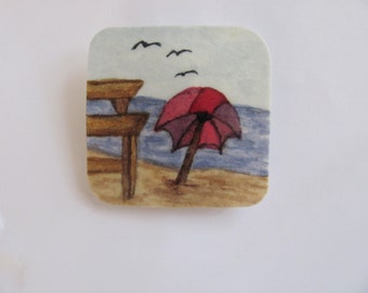 Könnte beinhalten: Ein Aquarellbild, das eine Strandszene mit einem roten und violetten Sonnenschirm, einer Holzbank und einem blauen Meer zeigt.