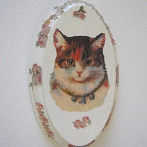 May include: A white oval-shaped ceramic plaque with a floral border and a vintage image of a cat with brown and white fur wearing a collar with a bell.