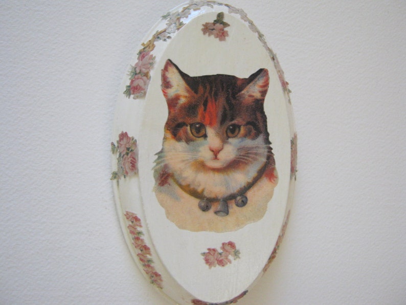 May include: A white oval-shaped ceramic plaque with a floral border and a vintage image of a cat with brown and white fur wearing a collar with a bell.