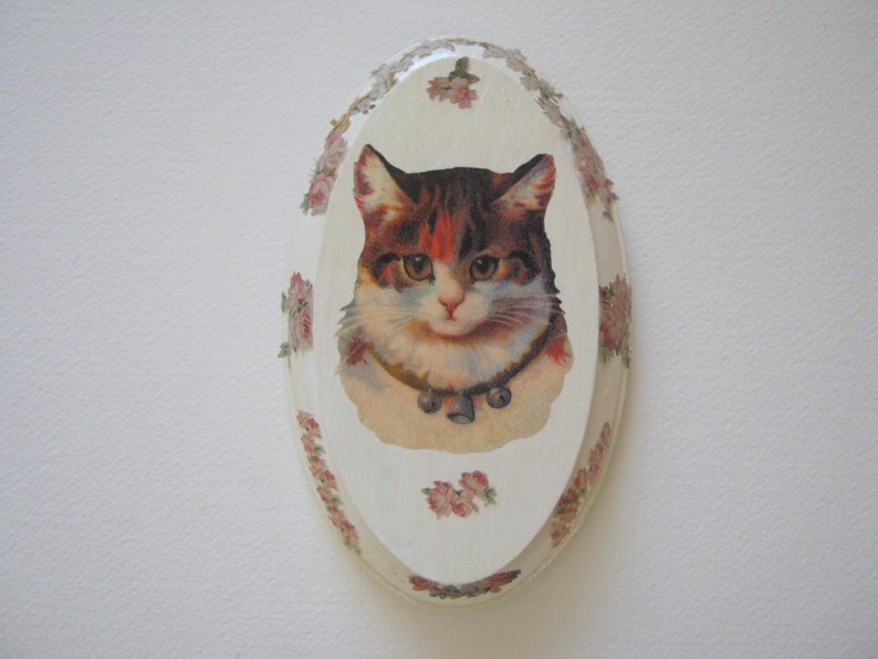 May include: A white oval-shaped ceramic trinket box with a floral design and a picture of a cat with a collar.