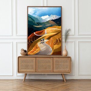 May include: A framed print of a colourful abstract landscape with two cyclists riding up a winding road. The road is surrounded by hills and mountains with a blue sky and white clouds in the background.