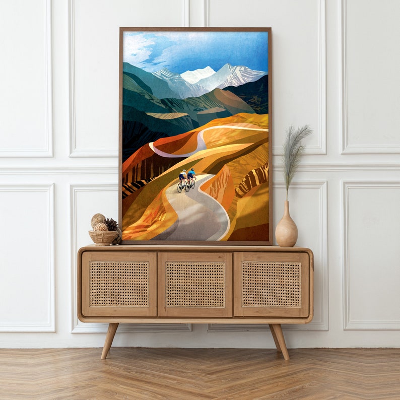May include: A framed print of a colourful abstract landscape with two cyclists riding up a winding road. The road is surrounded by hills and mountains with a blue sky and white clouds in the background.