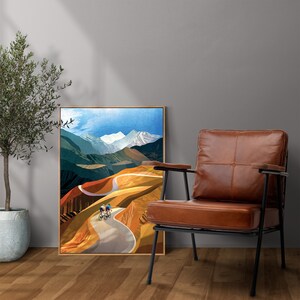May include: A framed print of a colourful landscape painting featuring a winding road through a mountain range with three cyclists riding up the hill. The print is hanging on a wall next to a brown leather armchair.