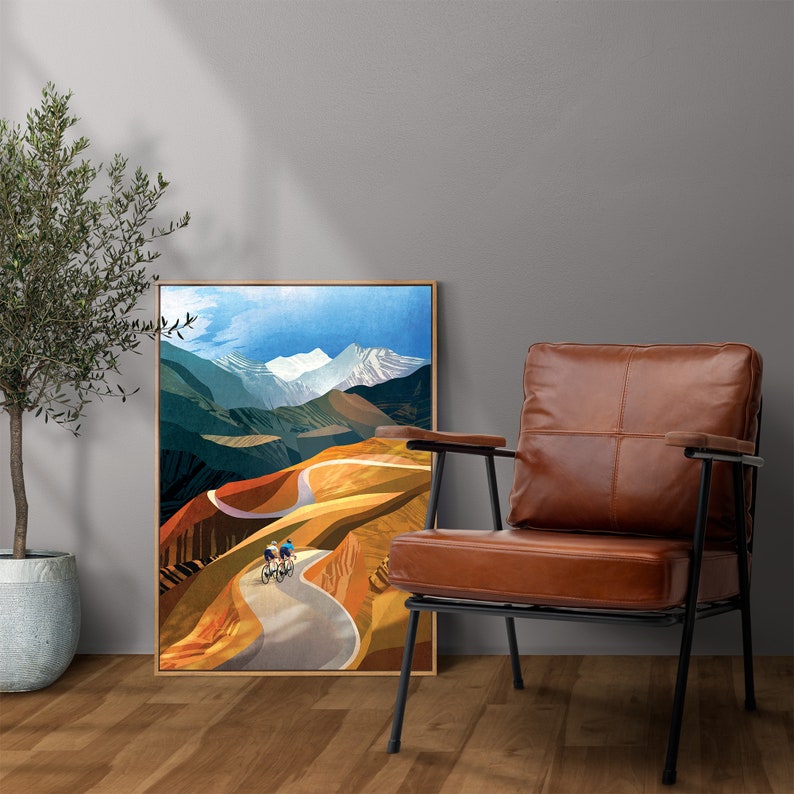 May include: A framed print of a colourful landscape painting featuring a winding road through a mountain range with three cyclists riding up the hill. The print is hanging on a wall next to a brown leather armchair.
