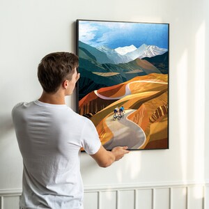 May include: A framed print depicting a colourful landscape with two cyclists riding on a winding road. The landscape is a mix of green, brown and orange hues, with a snowy mountain peak in the distance.