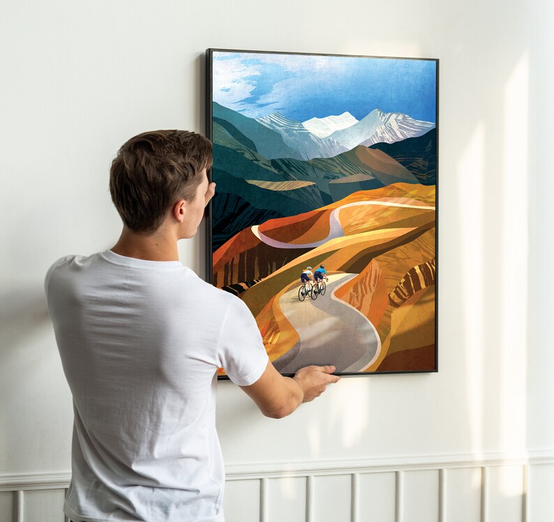May include: A framed print depicting a colourful landscape with two cyclists riding on a winding road. The landscape is a mix of green, brown and orange hues, with a snowy mountain peak in the distance.