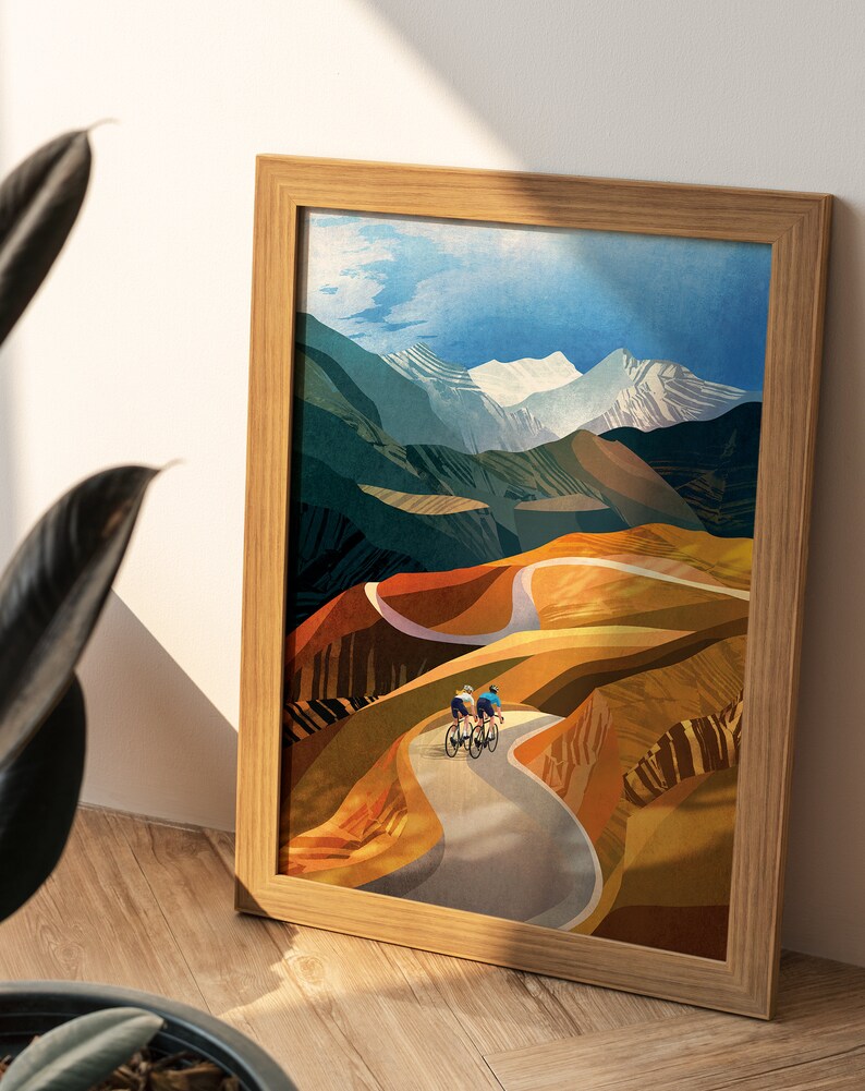 May include: Framed illustration of three cyclists riding on a winding road through a mountainous landscape. The mountains are painted in shades of blue, green, and brown, with snow-capped peaks. The road is paved and curves through the landscape. The cyclists are wearing colourful clothing and riding road bikes.
