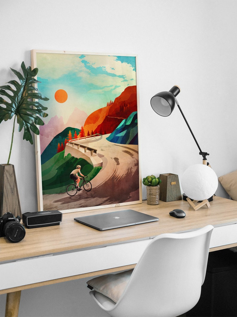 Pu&ograve; includere: Un poster incorniciato con un'illustrazione colorata di un ciclista che sale su una strada di montagna tortuosa. Il cielo &egrave; blu con nuvole bianche e un sole arancione brillante. Le montagne sono verdi, rosse e marroni.
