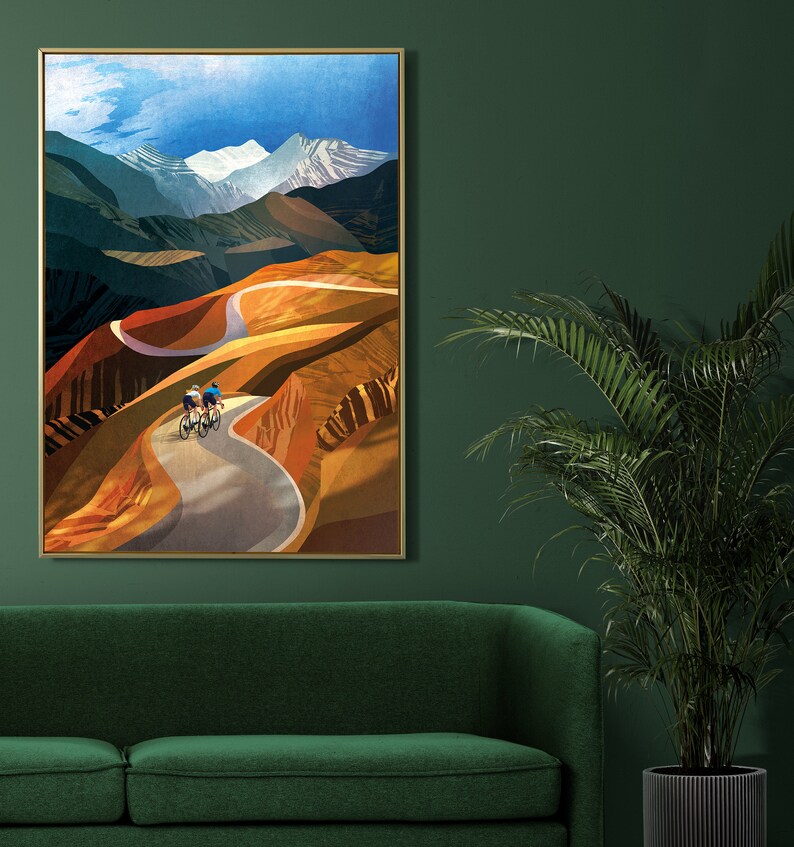May include: A framed print of a colourful landscape painting featuring two cyclists riding up a winding road through a mountain pass. The sky is blue with white clouds and the mountains are a mix of brown, green, and yellow.
