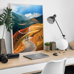 May include: A framed print of a colourful landscape painting featuring two cyclists riding on a winding road through a mountain range. The sky is blue with white clouds and the mountains are a mix of green, brown, and orange.
