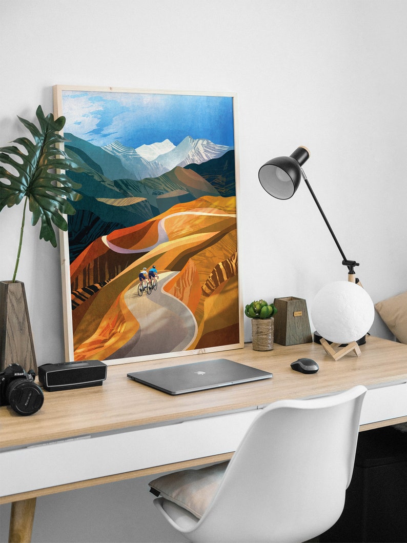 May include: A framed print of a colourful landscape painting featuring two cyclists riding on a winding road through a mountain range. The sky is blue with white clouds and the mountains are a mix of green, brown, and orange.