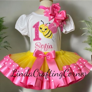 May include: A white short-sleeved shirt with a pink number 1 and the name "Sofia" is paired with a yellow tutu skirt with pink polka dot trim. A pink bow and a bee graphic complete the outfit. The outfit is for a child.