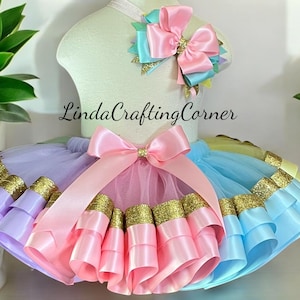 May include: A colorful tutu set with a matching headband, displayed on a mannequin. The tutu features layers of pink, purple, and blue fabric with gold trim. A large pink bow adorns the front, and the headband has a smaller bow.
