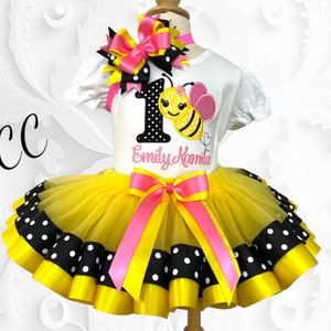 May include: Yellow and black polka dot tulle tutu with a pink and yellow ribbon bow. A white t-shirt with a bee and the number 1 embroidered on it. The shirt also has the name "Emily Kamala" embroidered on it.
