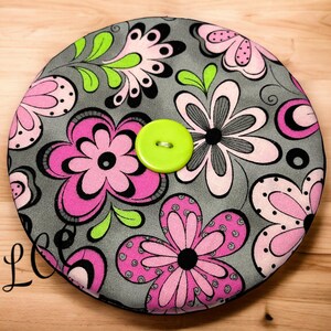 May include: A round gray fabric coaster with a floral pattern in pink, black, and green. The coaster has a lime green button in the center.