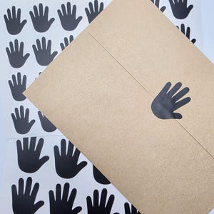 May include: A brown kraft paper envelope featuring a single black handprint sticker on the flap, positioned over a white sticker sheet containing multiple identical black handprint stickers arranged in a grid pattern across the surface.