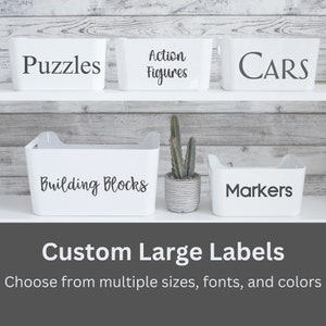 May include: Five white rectangular storage bins with integrated handles, each featuring black vinyl text labels reading Puzzles, Action Figures, CARS, Building Blocks, and Markers. A small gray ribbed planter with a green cactus sits between bins.