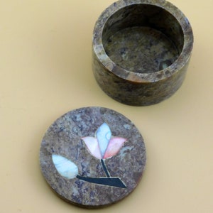 May include: A round, brown, stone trinket box with a lid featuring a flower design made of inlaid mother of pearl. The flower is pink, white, and blue.