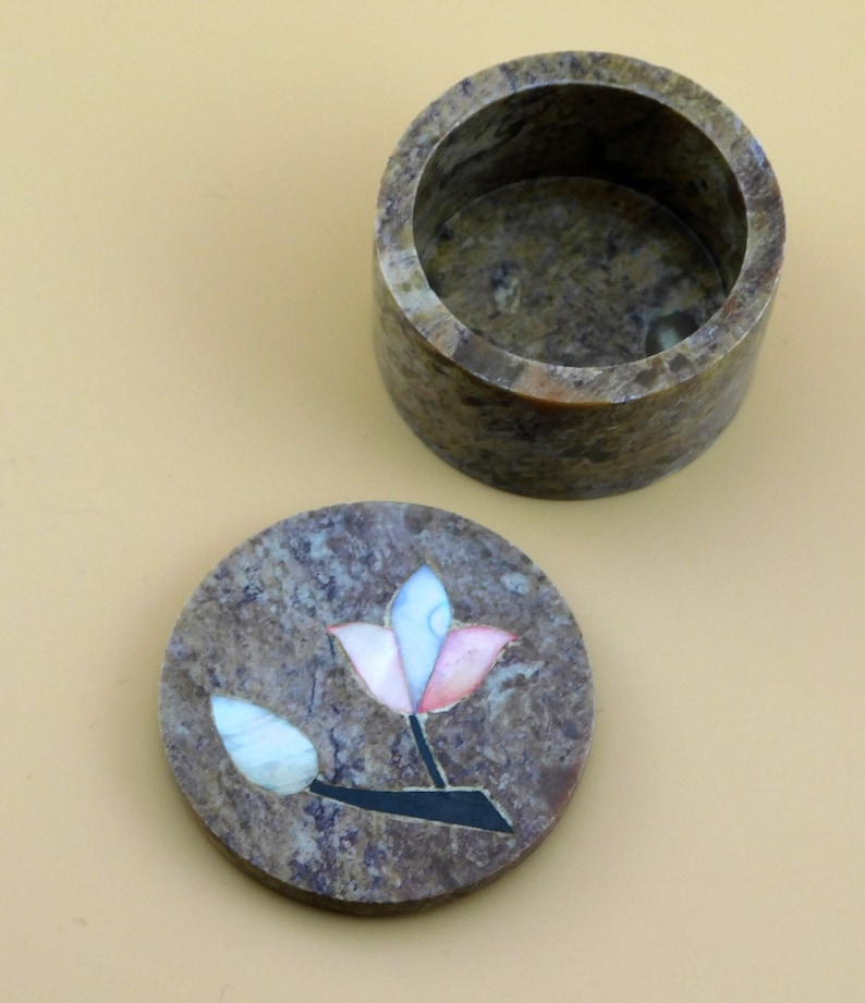 May include: A round, brown, stone trinket box with a lid featuring a flower design made of inlaid mother of pearl. The flower is pink, white, and blue.