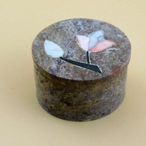 May include: A round, brown, stone trinket box with a floral design. The flower is made of inlaid pieces of white, pink, and blue stone.
