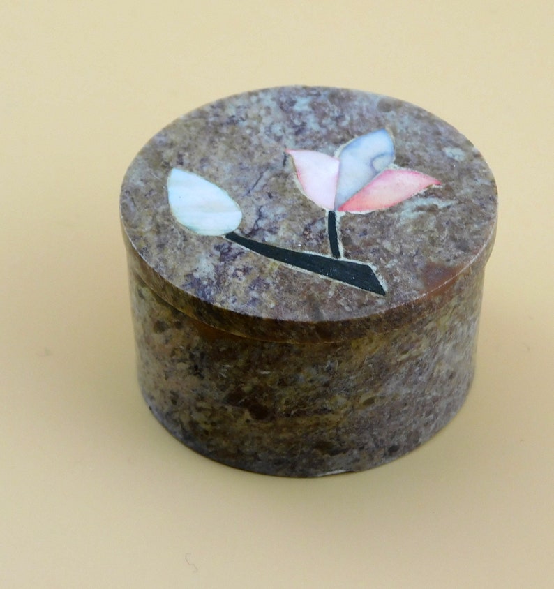 May include: A round, brown, stone trinket box with a floral design. The flower is made of inlaid pieces of white, pink, and blue stone.