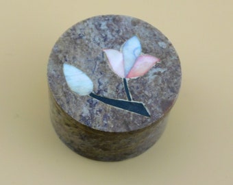 May include: A round, brown stone box with a floral design. The flower is made of inlaid pieces of colorful stone, including pink, blue, and white.